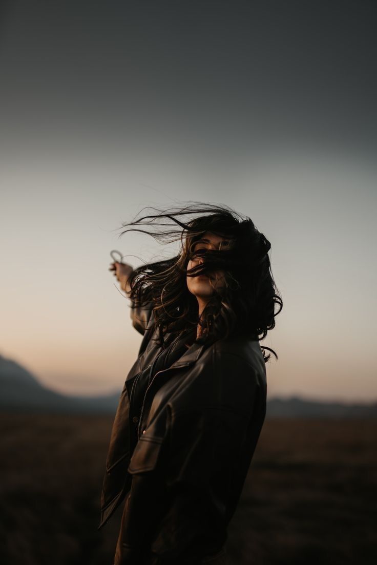 A woman in a leather jacket at dusk, her dark hair blown across her face, a soft orange horizon behind her.