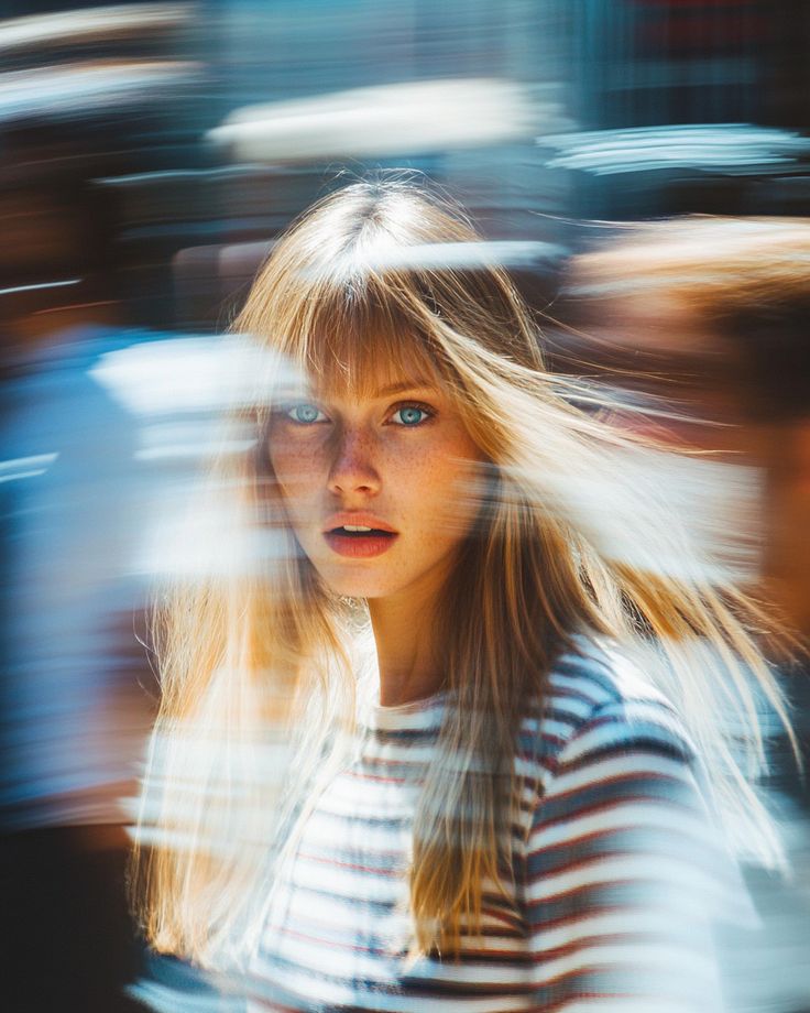 A young woman with long blonde hair and freckles looks straight into the lens while the street around her smears into streaks.