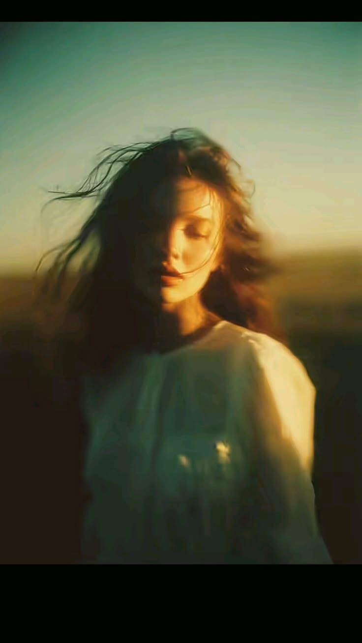A young woman in a white blouse at sunset, her hair blown across her face, the whole frame soft and golden.