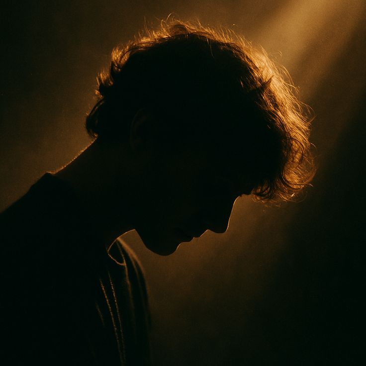 A young man in profile with his head bowed, his curly hair lit gold by a beam of light in a dark room.