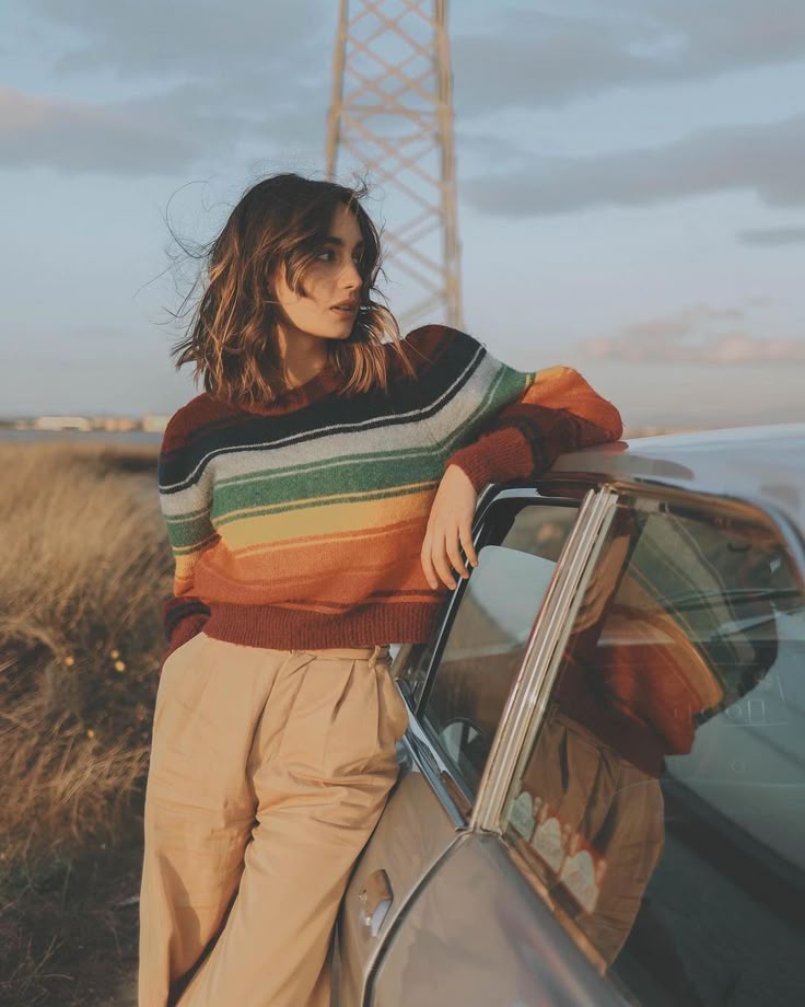 A young woman in a striped wool sweater leans on a car in golden evening light, her hair lifted by the wind.