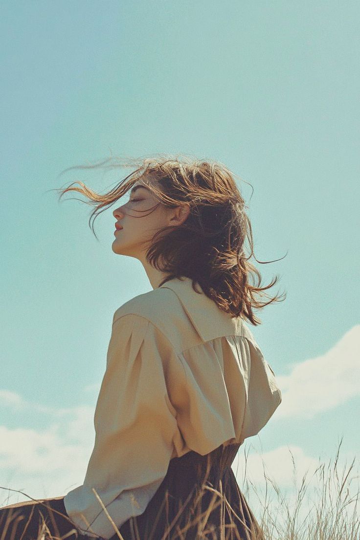 A woman with windswept auburn hair and a cream blouse turns her face to the sun against a pale blue sky.