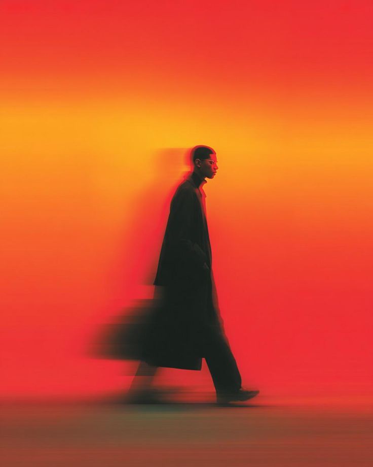 A man in a long dark coat walks across a glowing orange and red backdrop, his body trailing a motion blur.