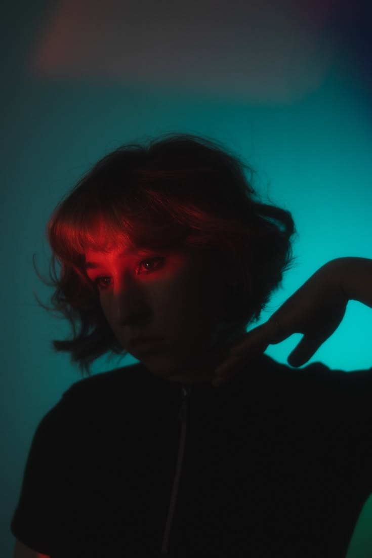 A young woman with a short bob, a red light across her eyes, against a glowing teal wall.