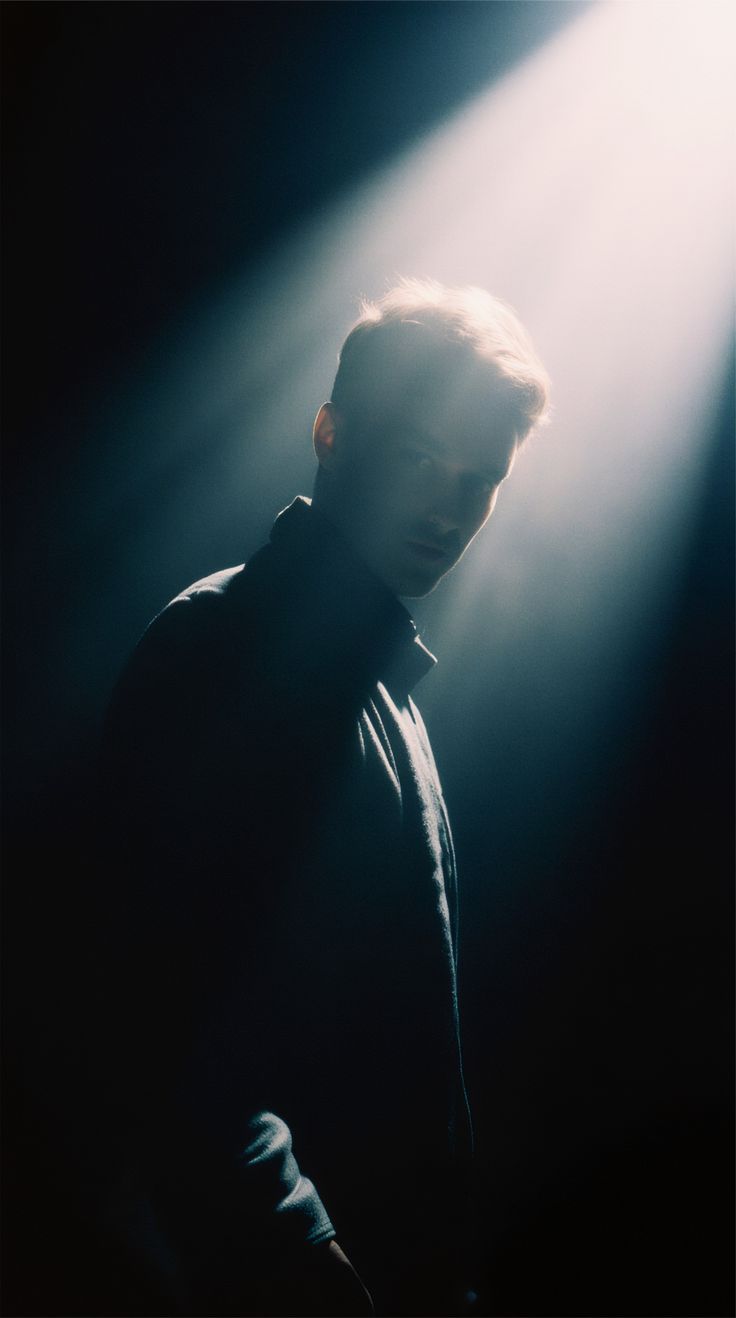 A man in a dark jacket stands in a shaft of white light that falls across his face from above.